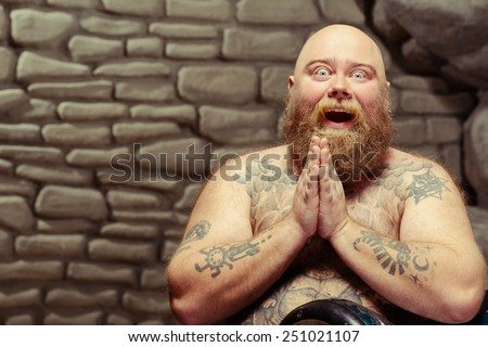 Wow. Portrait of funny bearded shirtless man with tattooed body playing the drum and keeping his palms together at his chin while sitting against stonewall background