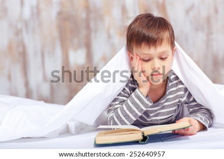 Intelligent little bookworm. Portrait of an adorable little boy lying in bed and reading a book with a blanket on his head