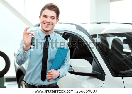 Key from your new car. Portrait of handsome young car sales man in formalwear holding a key from the car and smiling at camera in a car dealership