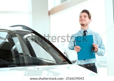 Perfect car dealer. Portrait of handsome young car sales man in formalwear holding a clipboard and looking up while standing by a new suv in a car dealership
