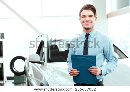 Perfect car dealer. Portrait of handsome young car sales man in formalwear holding a clipboard and smiling at camera in a car dealership