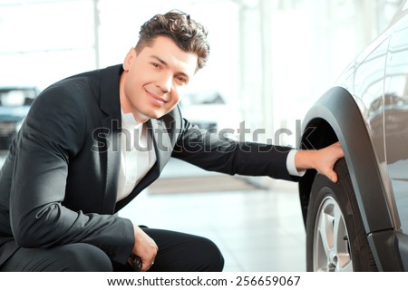 Confident in his choice. Smiling handsome man in suit checking wheels in a new car and smiling at camera in car dealership