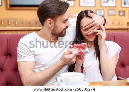 Surprise. Closeup of handsome young man giving his girlfriend a gift box and covering her eyes with hand while sitting in the cafe