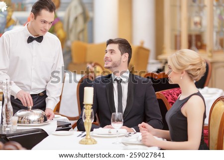 Couple in restaurant. Confident waiter serving the table with salad while beautiful couple looking at him and smiling while sitting in luxury restaurant with selective focus