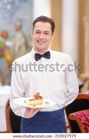 Try our tasty specials. Handsome waiter in uniform stretching out exquisite dessert  on the plate while serving customers in the luxury restaurant