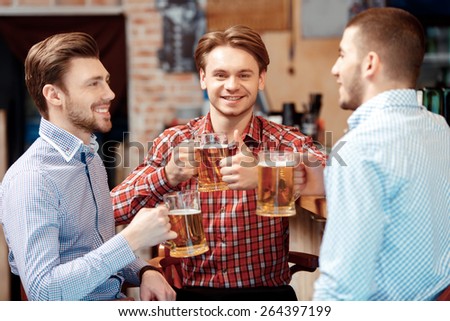 Cheers to friends. Three handsome young men in casual wear talking to each other and smiling while holding glasses with beer
