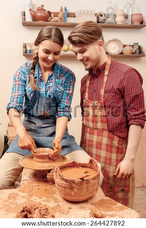 Pleasant work together.  Young female and male potters smiling and shaping on clay pot on the wheel in studio