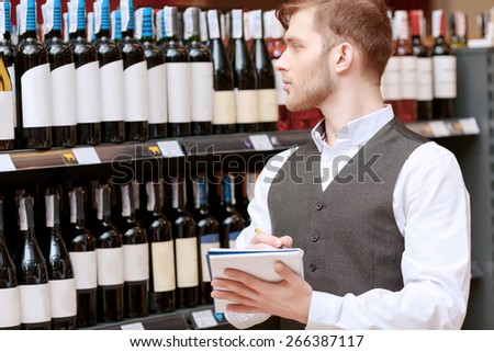 Young sommelier in a waistcoat making notes in his note pad standing by wine shelves and looking at the labels on bottles