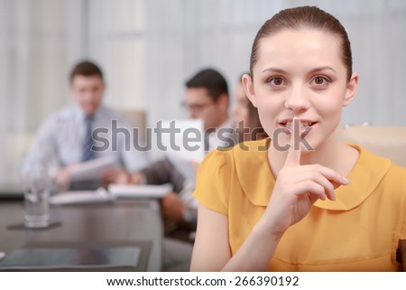 Keep it a secret. Young beautiful businesswoman showing at a camera sign to keep silent while her partners sitting in a row around the table in blurry
