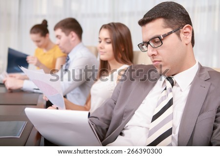 Need to check details. Confident strict business professional in formal wear and glasses reviewing business data at the meeting with his colleagues sitting in a row around the table