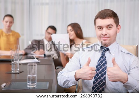 Best solutions for your business. Confident young businessman showing sign thumbs up at the business meeting with his team on the background