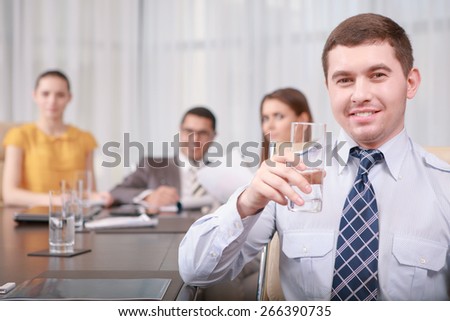 Have a sip of water. Young smiling manager wearing shirt and tie holding a glass of water at the business meeting with his team sitting on the background