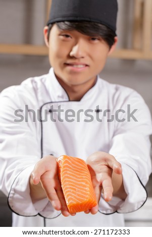 Only freshest fish. Young smiling Japanese cook stretching out to the camera piece of salmon with selective focus on fish