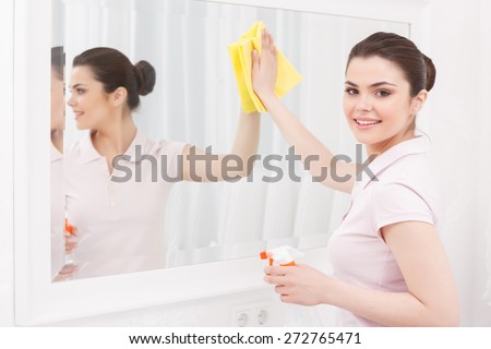 Cleanness in every move. Pretty brunette cleans mirror with help of yellow cloth and special mean of cleaning on white background