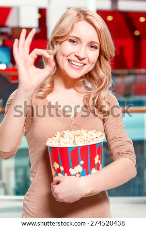 Everything is OK. Young blond woman standing in hall before watching film in cinema with popcorn showing OK.