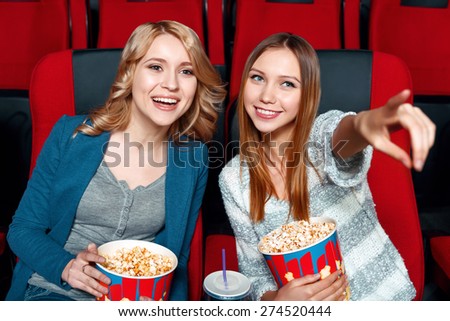 Look there. Two pretty smiling girls in cinema with popcorn and coke, one of them pointing straight.