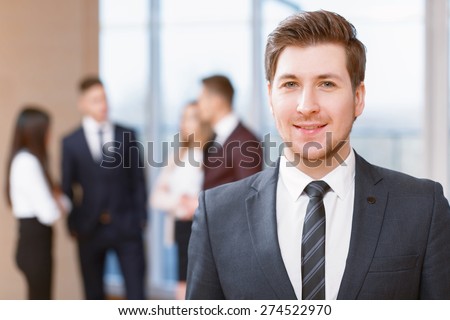 Office workers.  Young business man standing in foreground smiling, his co-workers discussing business matters in the background