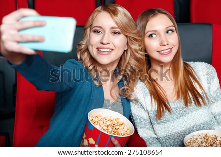 Cinema selfie. Two beautiful smiling girls excited doing selsie in cinema with popcorn and coke.