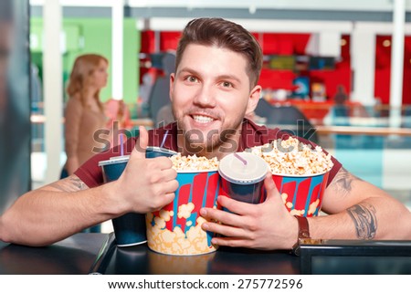 Greedy man. Young smiling friendly man buying popcorn and coke before watching film in cinema and showing class.