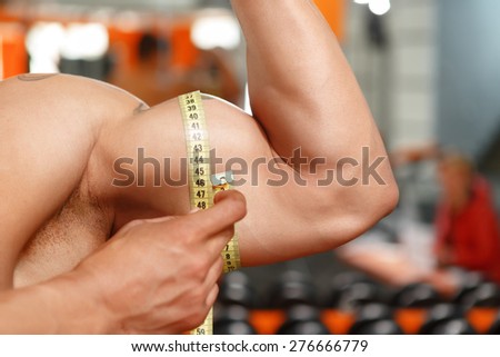 What size. Man measuring size of his biceps with help of soft tape measure in gym.