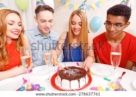 Birthday party. Young beautiful blond girl wearing cone cap cutting a birthday cake into pieces while her friends waiting and looking at it with appetite