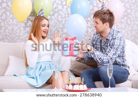 Young man presenting a birthday gift to his girl friend looking amused sitting on a couch wearing cone caps celebrating in a decorated room, cake standing on a table