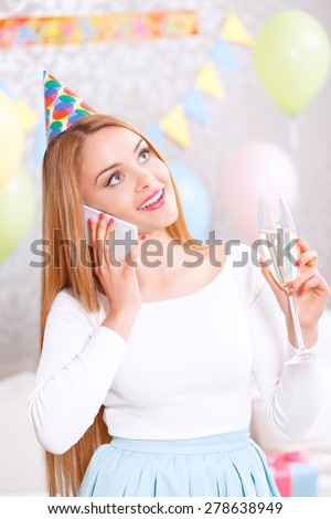 Birthday call. Young beautiful blond girl wearing cone cap taking congratulations on her birthday talking on her mobile phone and holding glass of champagne