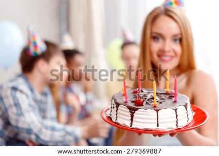 Sweetness. Portrait of a young beautiful blond girl wearing cone cap holding her birthday cake and her friends on the background celebrating