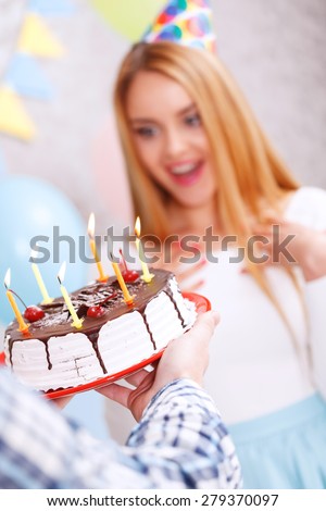 Is that for me. Young beautiful blond girl wearing cone cap looking surprised at birthday cake her friend holding in front of her