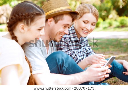 Having fun. Handsome guy and two pretty girls on picnic smiling, sitting on cover and looking on mobile phone.
