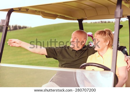 Look there. Senior smiling man and woman driving in cart on course, man pointing aside with help of his hand.