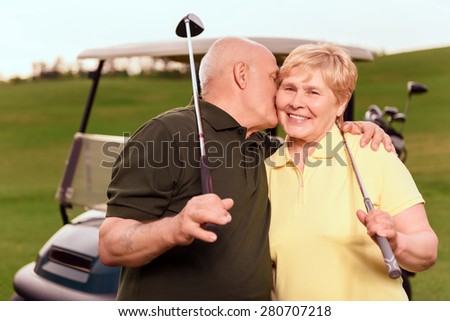 Showing love. Senior man with golf club giving kiss on cheek to his wife on background of cart on course.