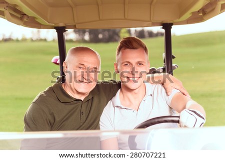 Supporting embrace. Smiling young and senior men driving in cart on course, old man embracing young one.