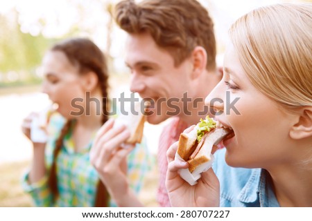 Biting off. Group of young people sitting on cover and eating sandwiches during picnic