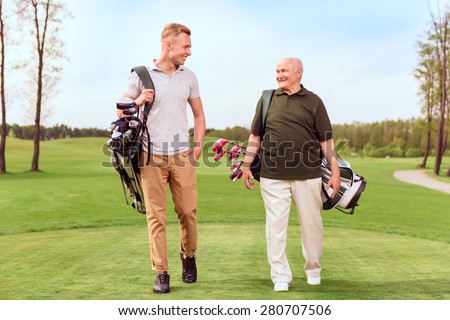 Two generations. Senior and young golf players walking through course with golf equipment.