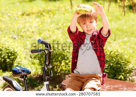 Look what I have. Little boy sitting on bench in park and holding hamburger above his head
