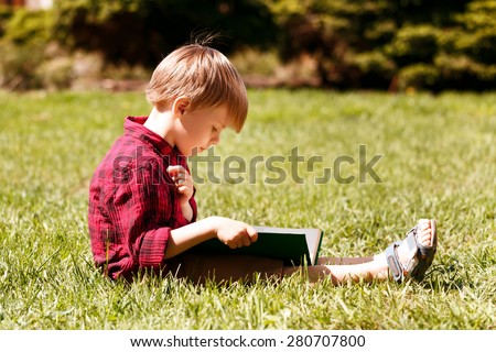Involved in reading. Portrait of little boy in profile sitting on grass and reading book