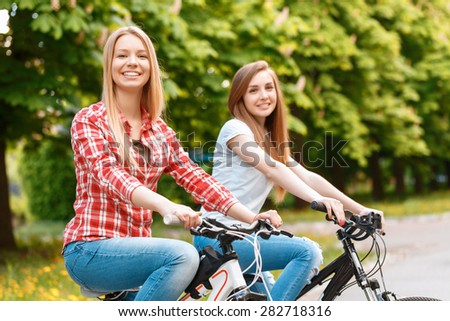 Two young beautiful girls with long hair smiling while riding their mountain bikes in a green park ,selective focus