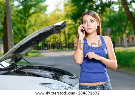 Felling shocked. Beautiful youthful lady standing on background of car with opened bonnet and using mobile phone.
