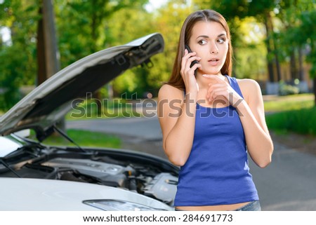 Feeling confused. Attractive young woman standing on background of car with opened bonnet and calling someone.