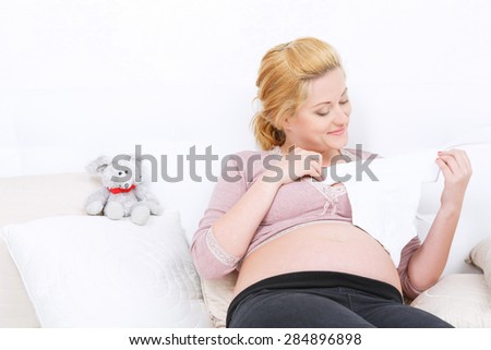Wishing girl. Smiling pregnant woman holding baby clothes and keeping eyes on it while lying on bed.