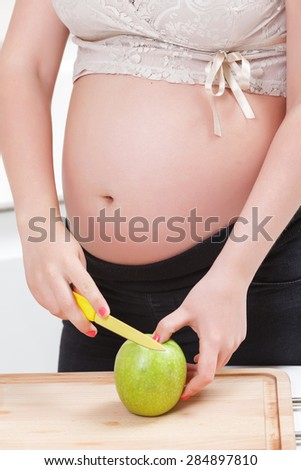 Healthy lifestyle. Pregnant young woman in cutting an apple holding it  on the wooden chopping board while standing in the kitchen.