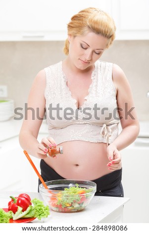 Tasty cooking. Pregnant young woman keeping eyes down and seasoning vegetable salad with spices while cooking in the kitchen.