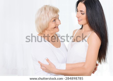 Soft glance. Pretty grandmother and granddaughter standing and embracing glancing at each other on white background
