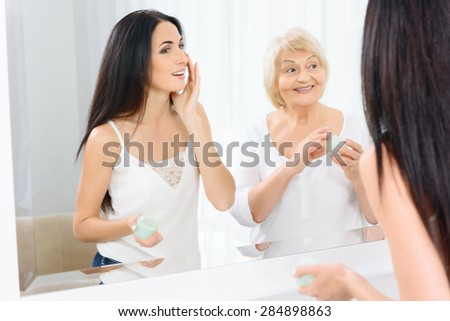 Taking care. Pair of two women standing in front of mirror and putting cream on their faces.
