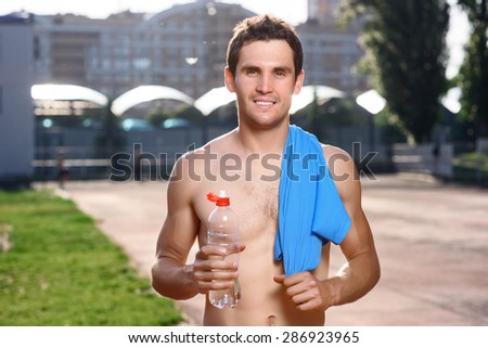 Water is life. Young smiling handsome topless man standing in stadium with bottle of water