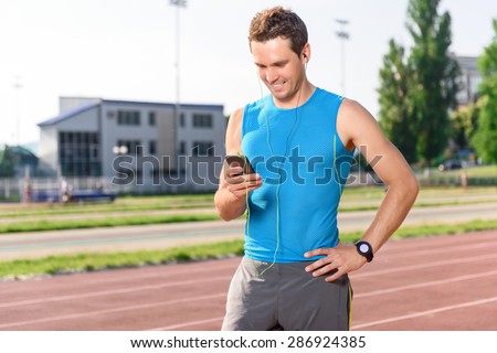Choosing track. Young handsome sportsman standing on stadium and holding mobile phone.