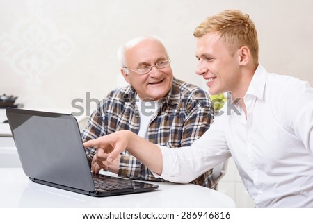 Pay attention. Grandson sitting together with his grandpa in kitchen and pointing on laptop