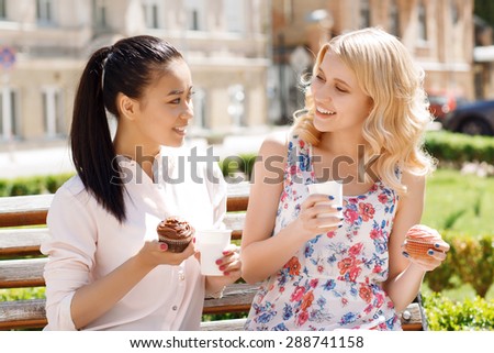 Funny conversation. Two young smiling girlfriends sitting in park and holding cupcakes with coffee.