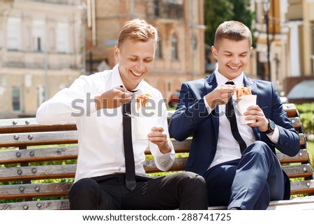 Pleasant moment. Pair of young handsome men in suits sitting on bench and eating Chinese noodles.